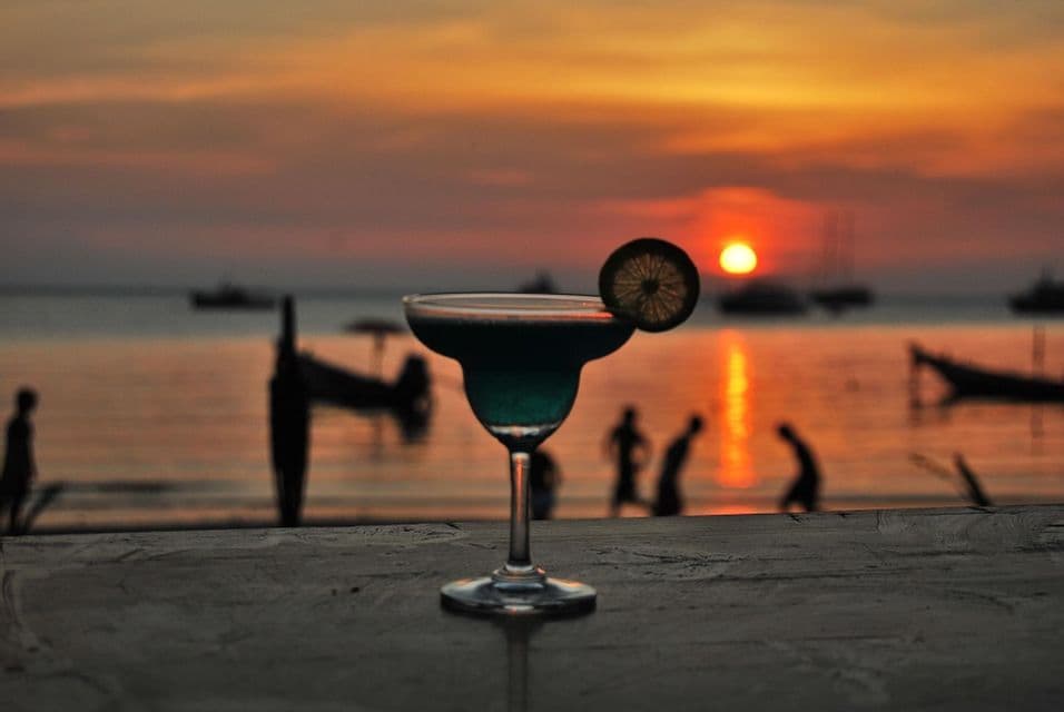 Un cocktail blu con guarnizione di lime su un tavolo, con vista su una spiaggia e sagome di persone e barche al tramonto.