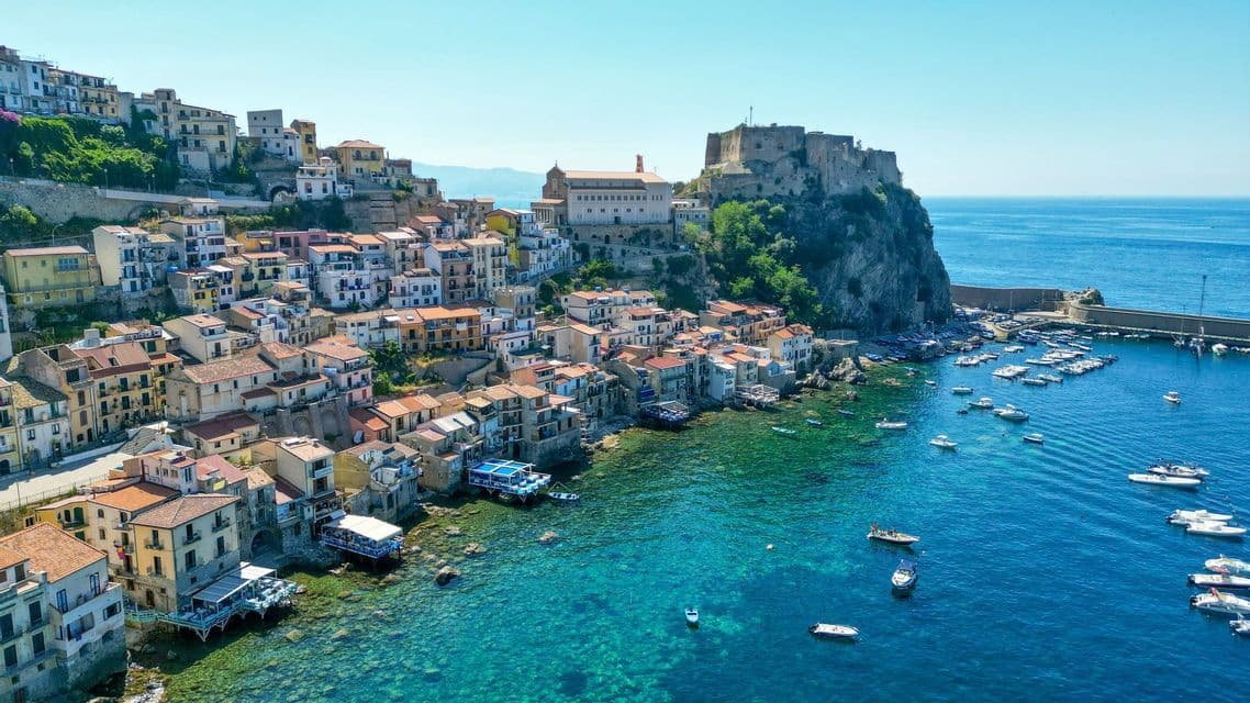 Una vista aerea di un vivace borgo costiero su una scogliera, con un castello e un porto pieno di barche in acque turchesi.