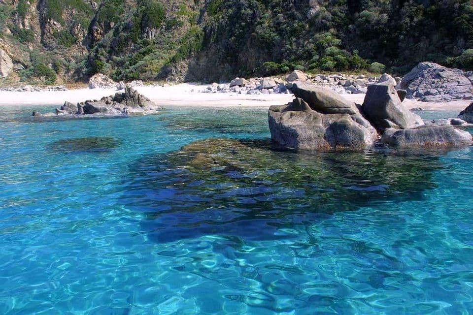 In una cala appartata, l'acqua turchese e limpida circonda grandi rocce, con una spiaggia di sabbia bianca e una scogliera verde e rocciosa a fare da sfondo.