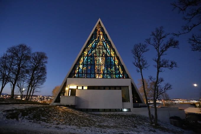 Una iglesia moderna y triangular con un gran vitral iluminado en una colina nevada al anochecer.