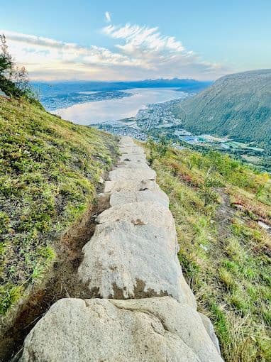 Un sendero de piedra desciende por una ladera montañosa verde, con vistas a una ciudad y un fiordo abajo.