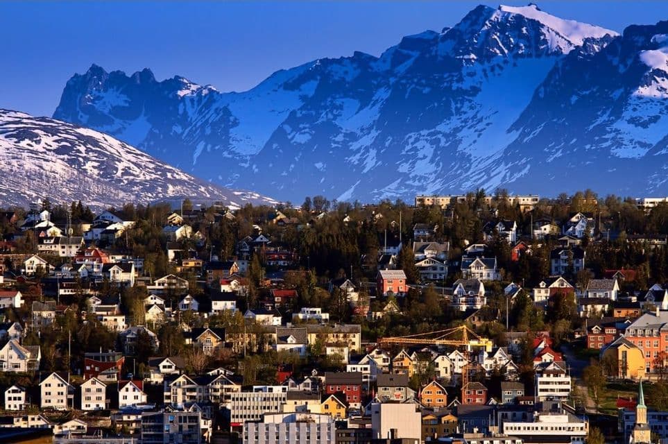 Un pueblo denso y colorido en una ladera se asienta al pie de grandes montañas nevadas bajo un cielo azul claro.