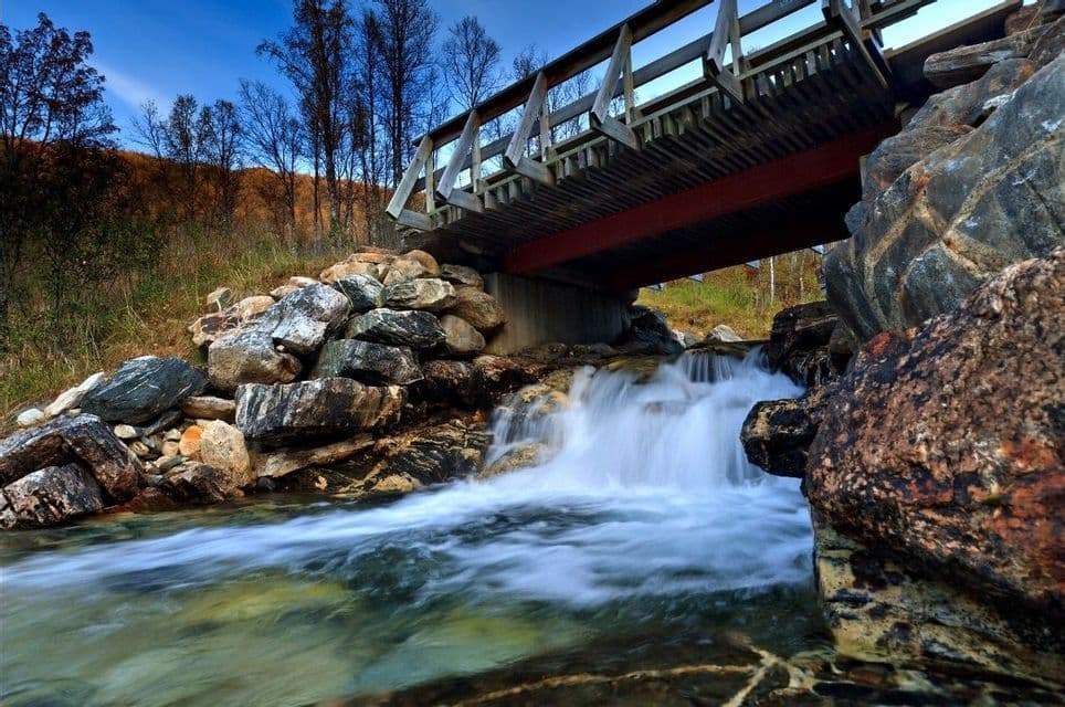 Una foto a lunga esposizione mostra un ruscello che scorre sulle rocce, creando una piccola cascata sotto un ponte di legno.