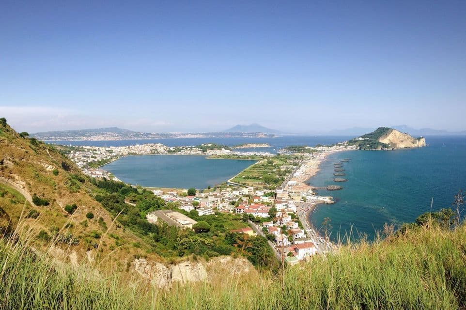 Una vista panorámica desde una colina cubierta de hierba de un pueblo costero que rodea una bahía, con una playa y montañas distantes bajo un cielo azul.