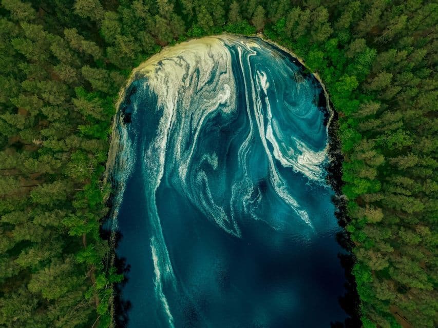 Vista aerea di un lago blu scuro, con motivi bianchi e gialli vorticosi sulla sua superficie, circondato da una fitta foresta verde.