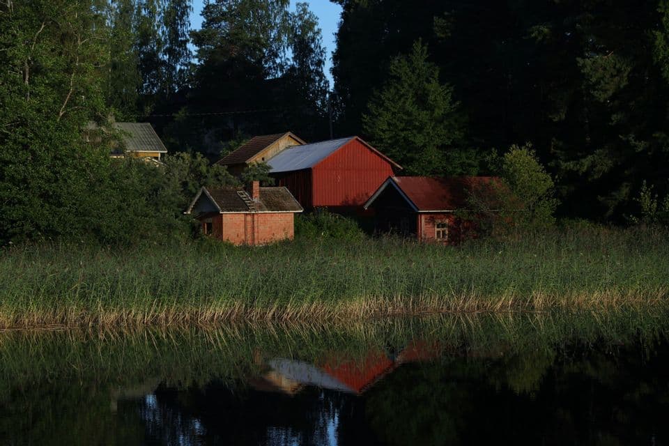Diversi edifici rustici, tra cui un fienile rosso, sono annidati tra gli alberi sulla riva di un lago e si riflettono nelle calme acque sottostanti.