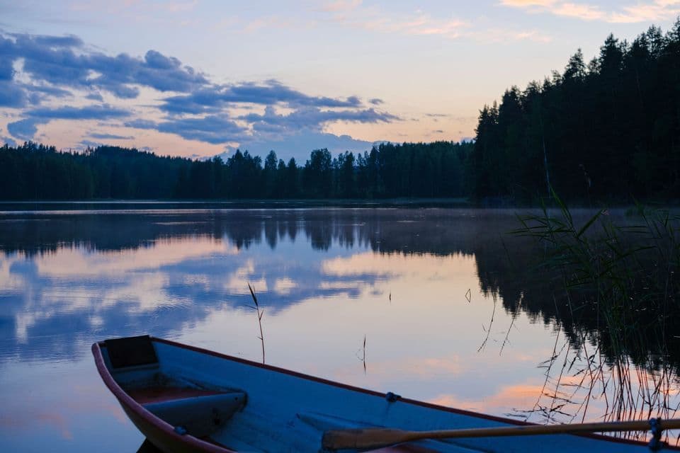 La prua di una piccola barca riposa sulla riva di un lago calmo al tramonto, con il cielo e una foresta riflessi sull'acqua.