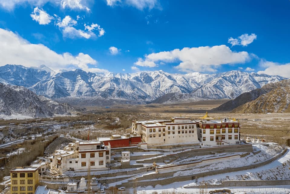 A large monastery complex sits on a snow-covered hillside with a vast, snow-capped mountain range in the background under a blue sky.