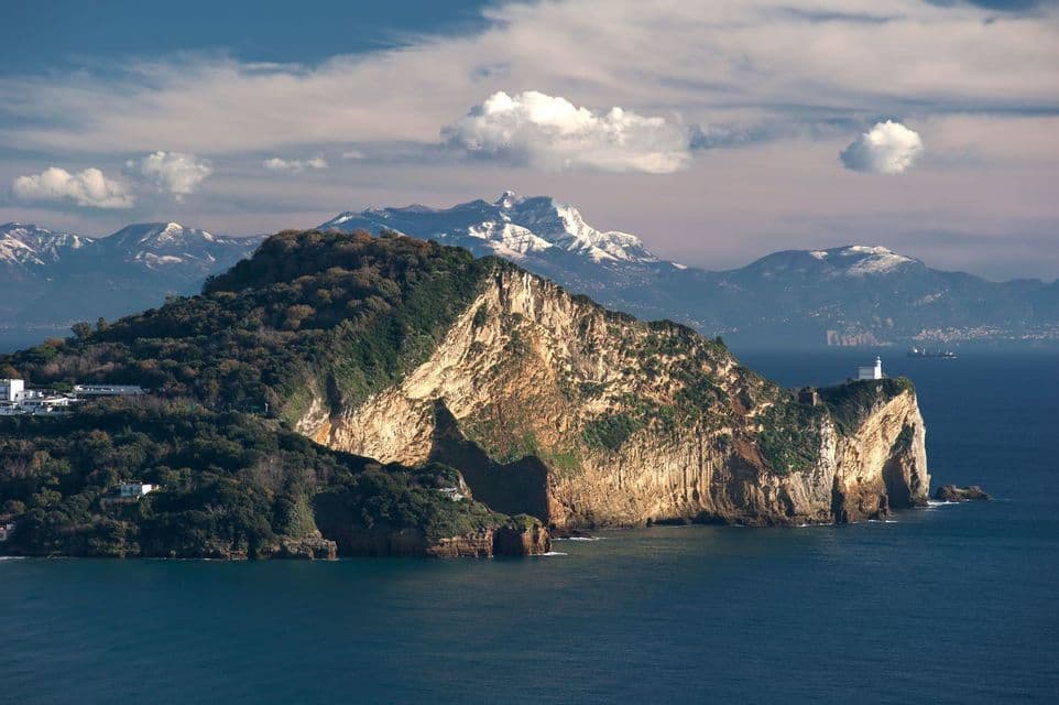Una isla rocosa cubierta de árboles que se adentra en el mar, con un faro en su punta y montañas nevadas al fondo.