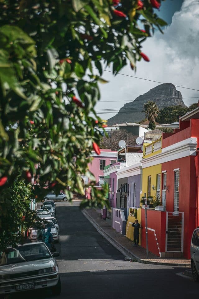 Une rue en pente bordée de maisons colorées en rouge, jaune et violet, avec une grande montagne visible en arrière-plan sous un ciel nuageux.