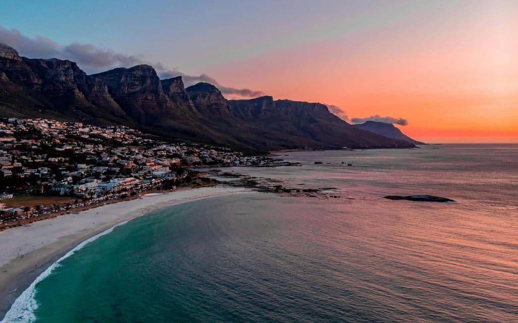 Une vue aérienne d'une ville côtière et d'une plage de sable nichées au pied d'une chaîne de montagnes, sous un coucher de soleil coloré.