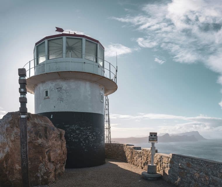 Un phare noir et blanc se dresse sur un promontoire rocheux dominant l'océan et les montagnes lointaines sous un ciel partiellement nuageux.