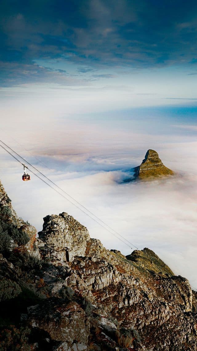 Un téléphérique descend une montagne rocailleuse, bien au-dessus d'une épaisse mer de nuages d'où émerge un sommet lointain.