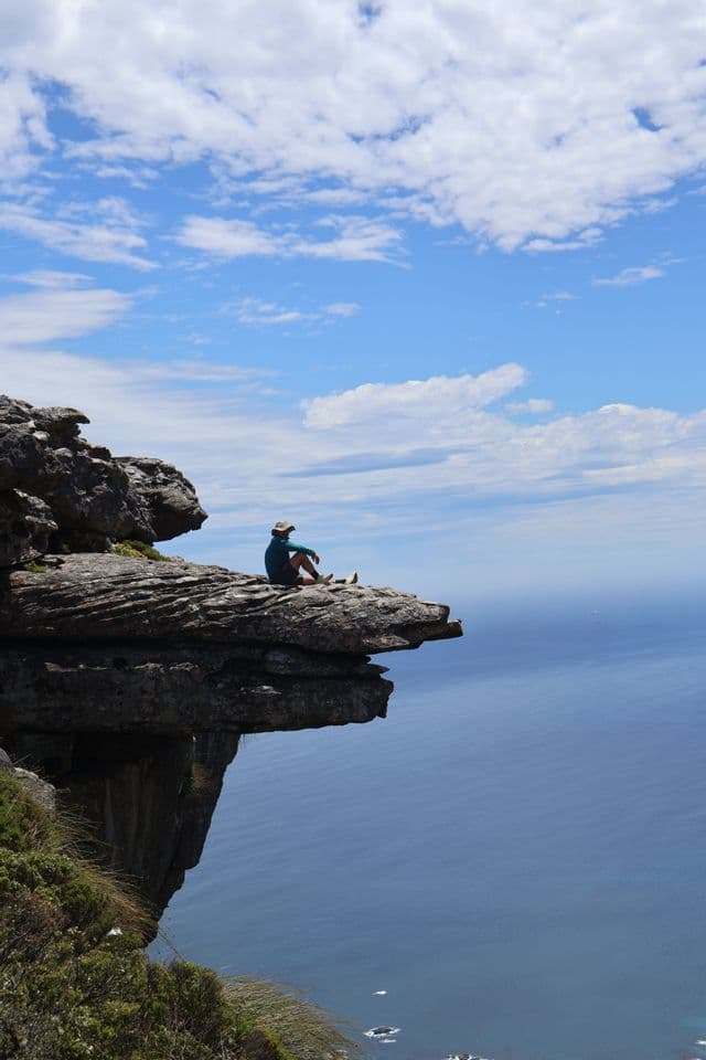 Une personne portant un chapeau est assise au bord d'une falaise rocheuse, contemplant le vaste océan bleu sous un ciel partiellement nuageux.