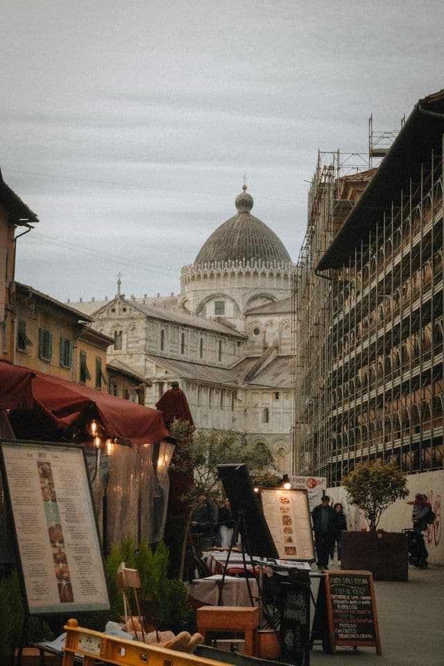 Blick auf eine enge Stadtstraße mit einer großen Kuppelkathedrale, Außengastronomie und einem Gebäude unter Gerüsten im Vordergrund.