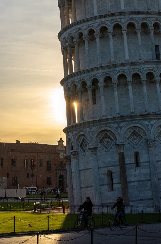 Zwei Personen radeln bei Sonnenuntergang an einem schiefen Steinturm vorbei, wobei die Sonne durch dessen Bögen scheint.