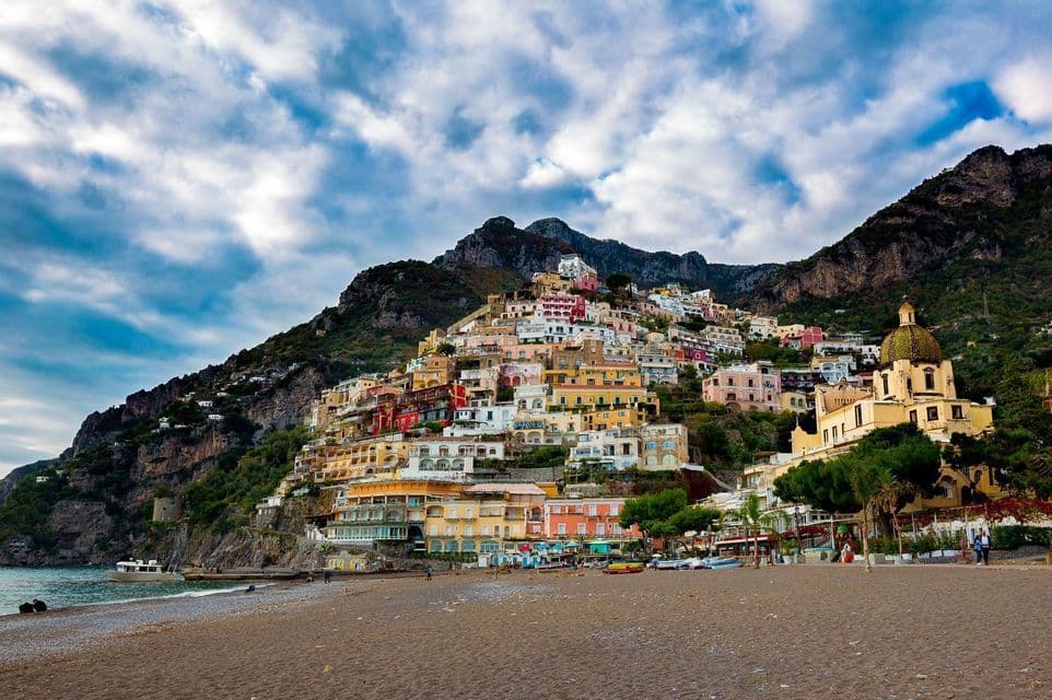 Una vista desde una playa de arena de un pueblo costero con casas coloridas construidas en una empinada ladera verde bajo un cielo dramático y nublado.