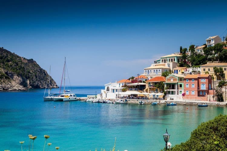 Barche a vela e un catamarano sono ormeggiati in una baia turchese di fronte a un colorato borgo marinaro su una collina.