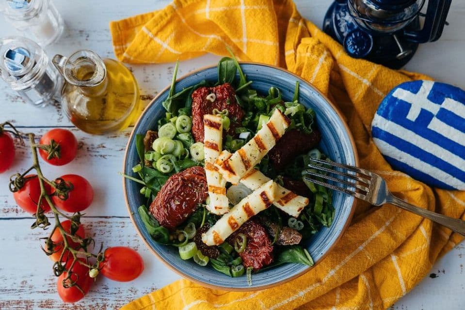 Una vista dall'alto di un'insalata con halloumi grigliato e pomodori secchi, accanto a una roccia dipinta con la bandiera greca.