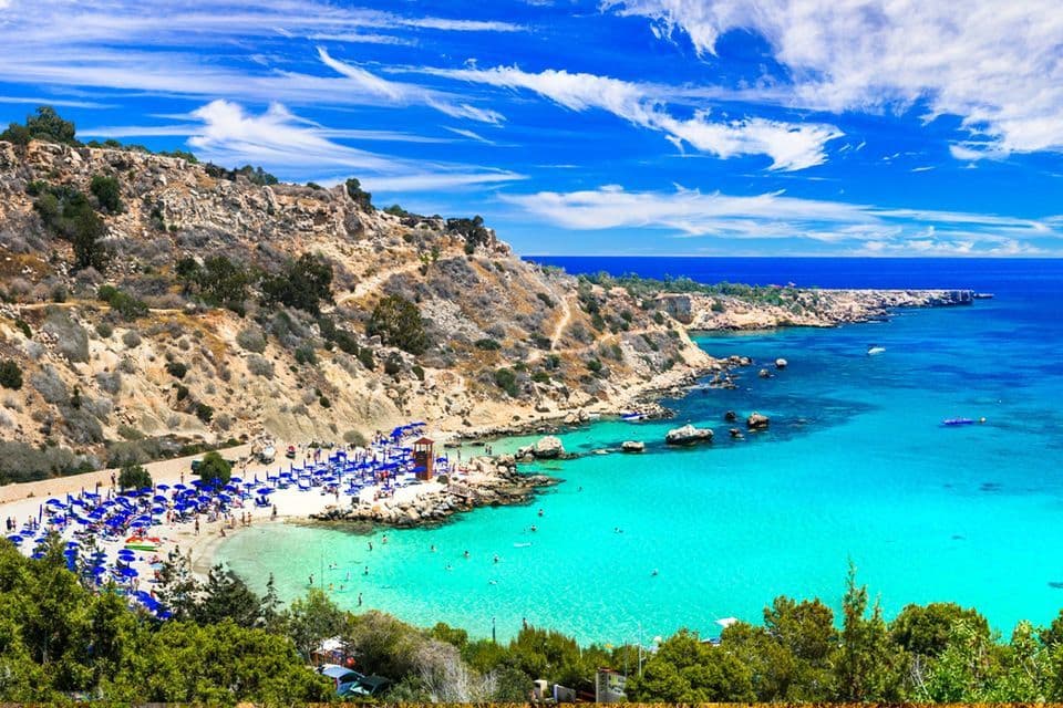 Una vista dall'alto di una caletta affollata con acqua turchese, ombrelloni blu e scogliere rocciose circostanti.