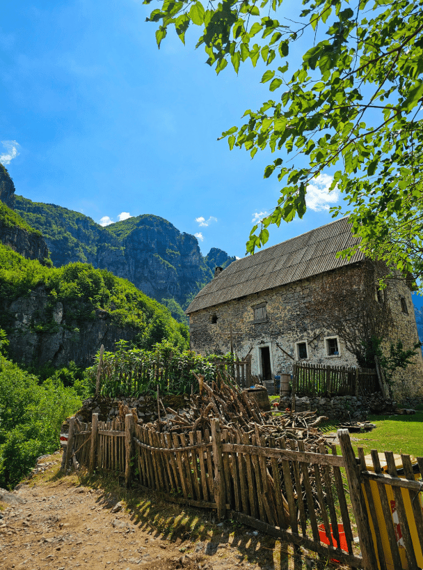 A rustic stone house sits on a green mountainside behind a wooden fence and a dirt path under a clear blue sky.