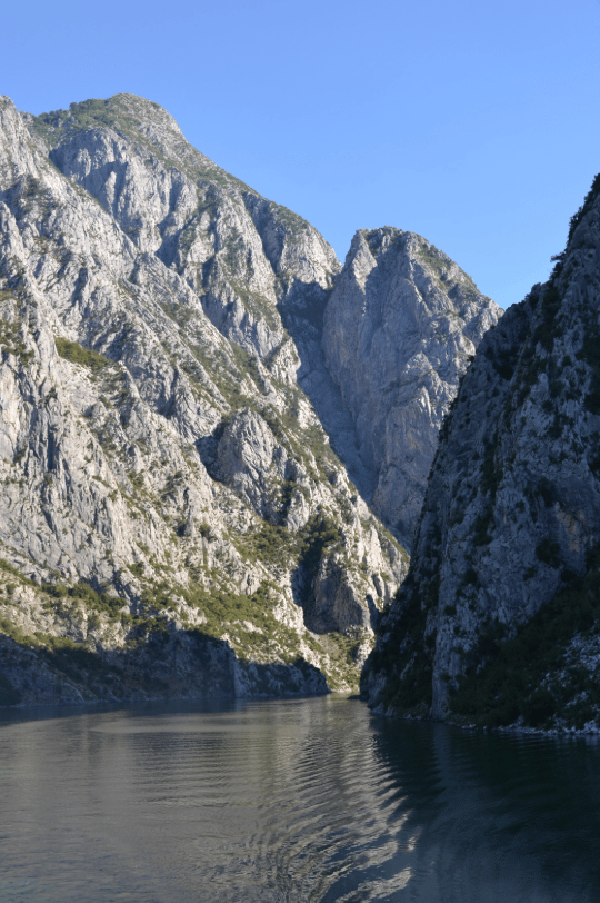 Steep, rocky mountains with sparse vegetation rise on either side of a calm river, forming a narrow canyon under a clear blue sky.