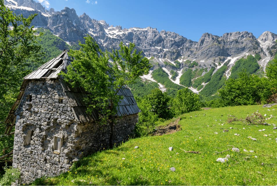 A rustic stone house with a damaged roof sits on a green hillside in front of a rugged mountain range with snow patches.