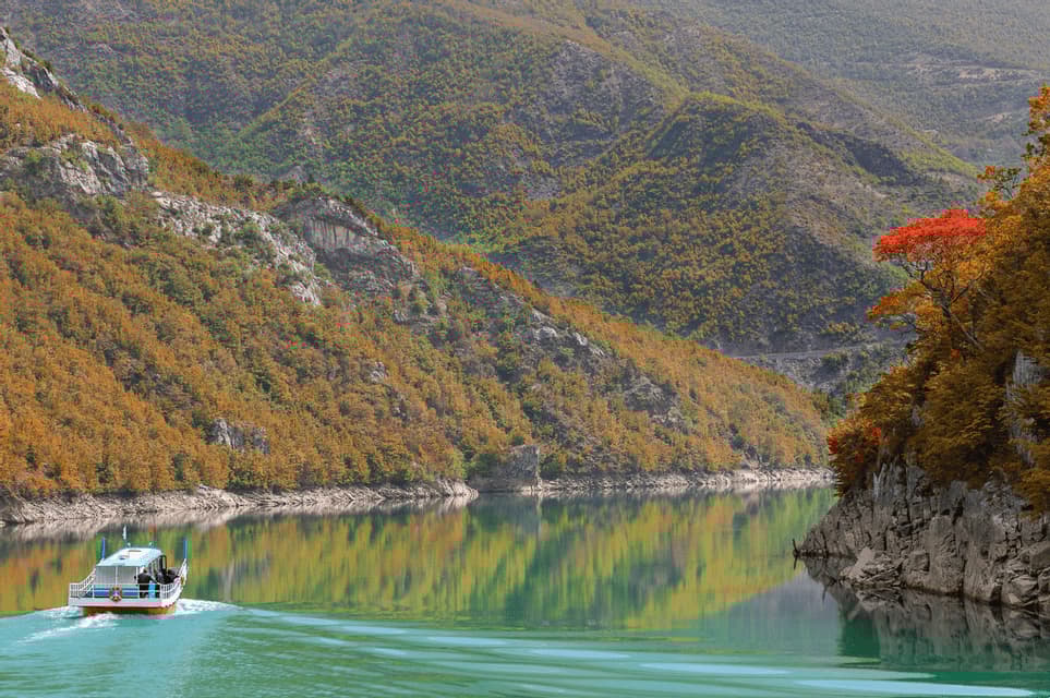 A WeRoad group trip on a tour boat sailing on a calm turquoise lake, surrounded by mountains with autumn foliage.