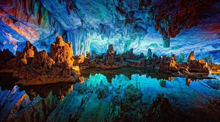 Les formations de stalactites et de stalagmites dans une grotte sont illuminées par des lumières multicolores, se reflétant parfaitement dans l'eau immobile.