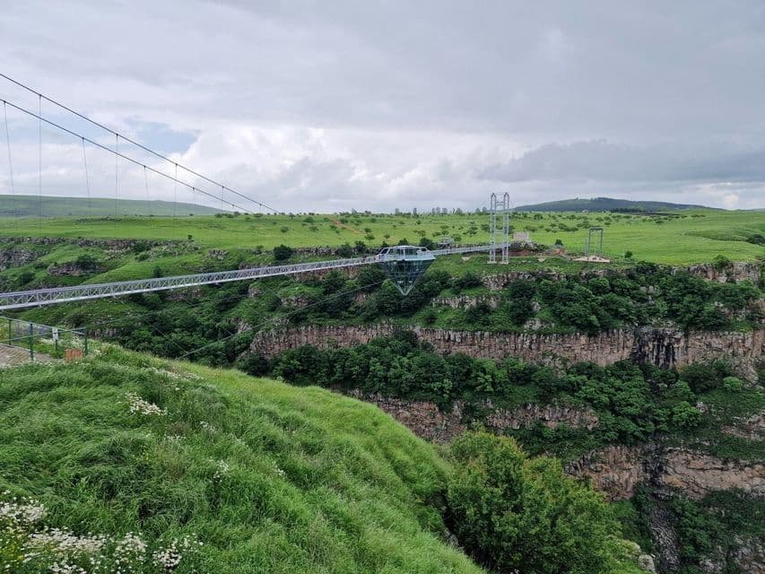 Un pont suspendu moderne doté d'une structure centrale en verre en forme de diamant enjambe un canyon profond, niché entre des collines verdoyantes et vallonnées.