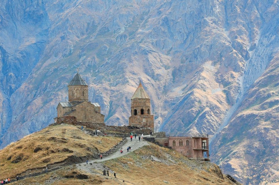 Des gens montent un sentier vers une église en pierre et un clocher sur une colline herbeuse, avec une vaste chaîne de montagnes accidentée en arrière-plan.