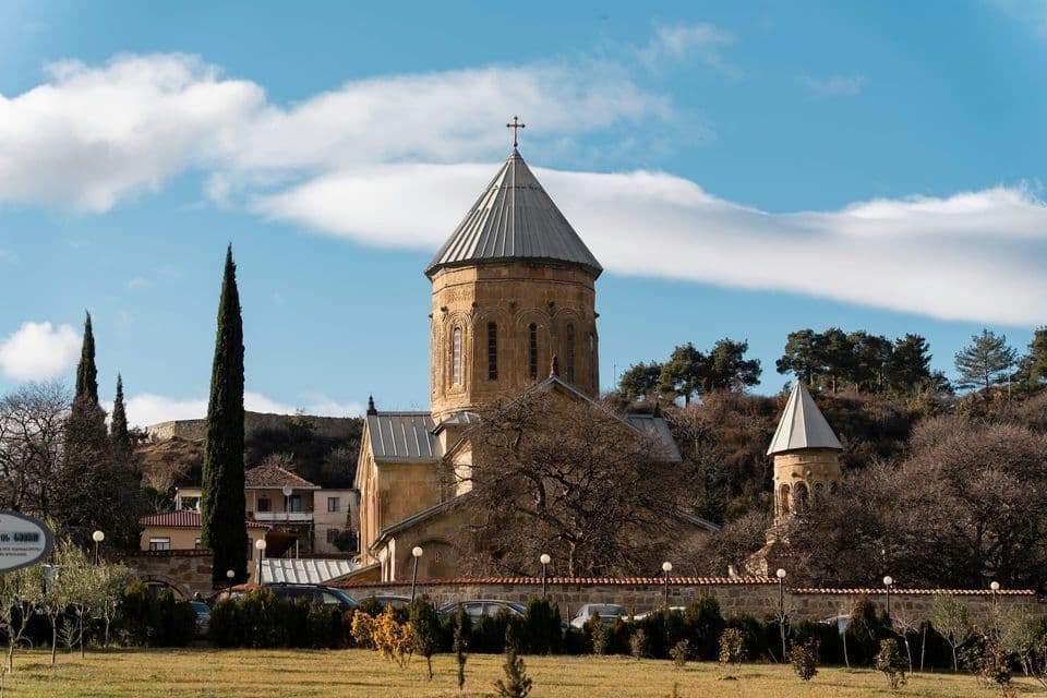 Une église en pierre au toit conique se dresse dans une zone herbeuse avec des arbres sous un ciel bleu partiellement nuageux.