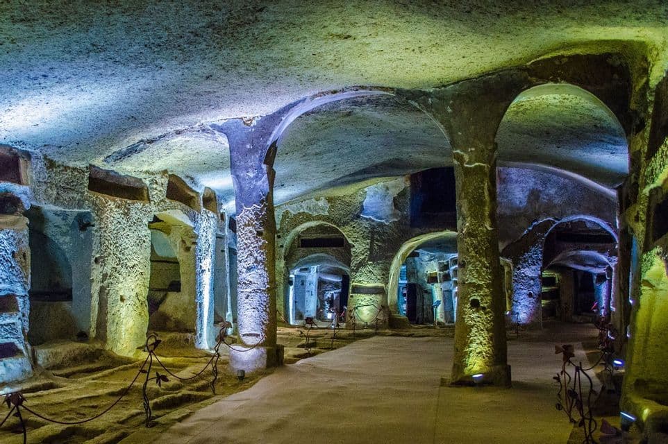Una gran catacumba subterránea con pilares y arcos de piedra toscamente tallados, iluminada por luces azules y amarillas.