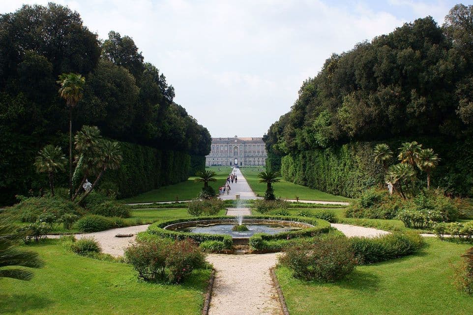 Un largo y simétrico camino de jardín flanqueado por altos árboles conduce a un gran palacio, con una fuente circular en primer plano.