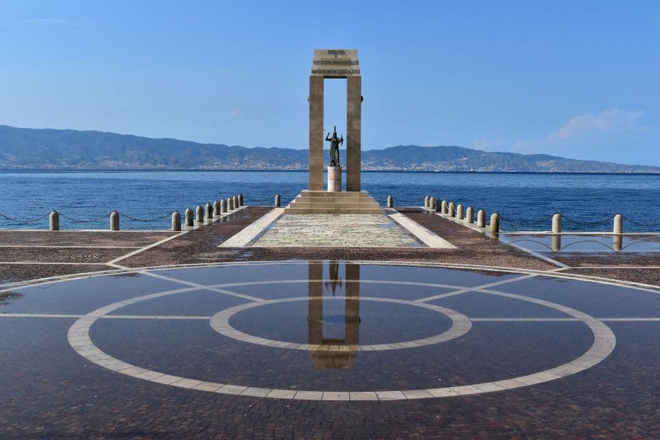 Un monumento su un molo si specchia in una vasca d'acqua circolare sulla pavimentazione, con il mare e le montagne sullo sfondo.