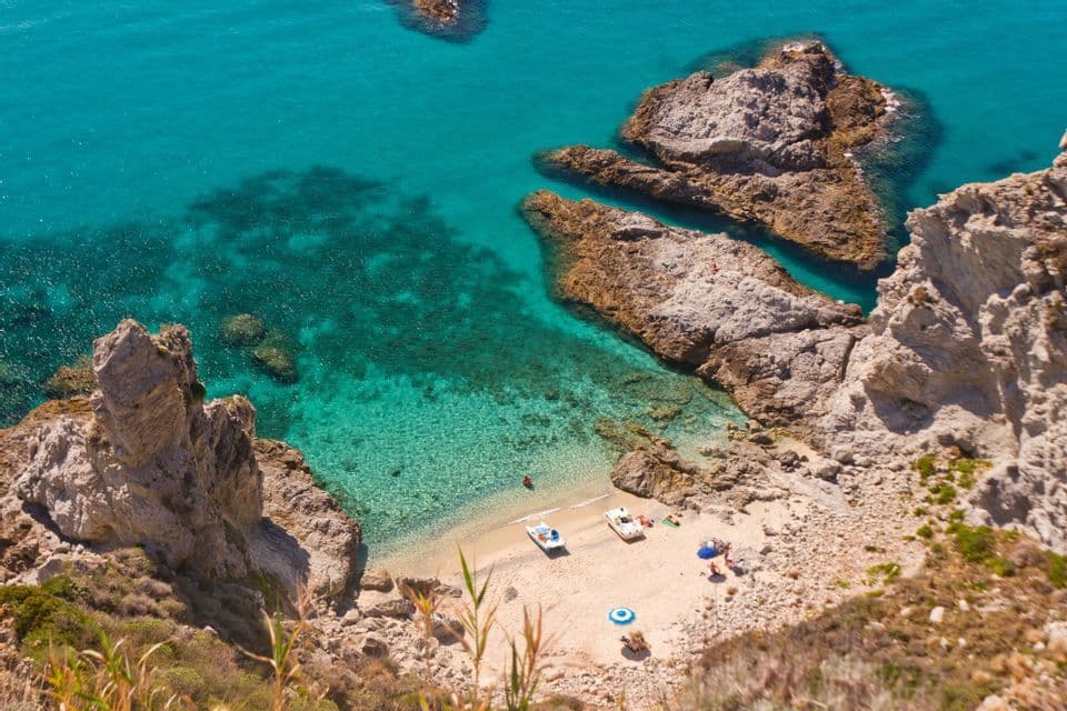 Una vista aerea di una spiaggia sabbiosa e isolata in una cala rocciosa, con acqua cristallina turchese e piccole barche a riva.