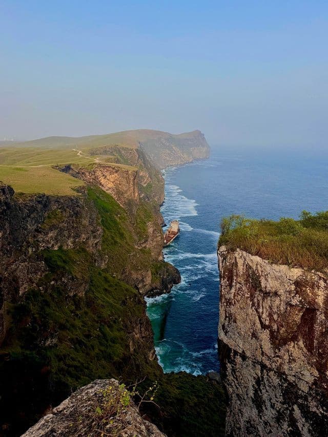 Una vista dalla scogliera su un relitto incagliato in una stretta insenatura tra ripide scogliere erbose e il mare blu.
