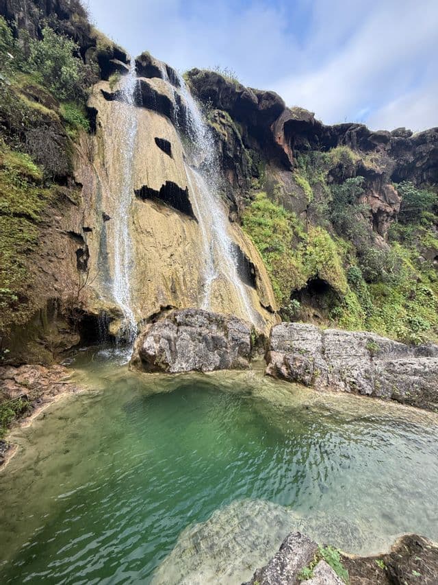 Una cascata scende lungo una parete rocciosa coperta di muschio in una piscina naturale limpida e verde sotto un cielo nuvoloso.