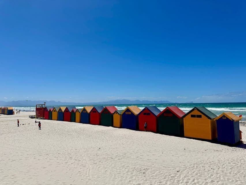Una lunga fila di cabine colorate fiancheggia una spiaggia sabbiosa, con l'oceano e le montagne distanti sotto un cielo azzurro e limpido.