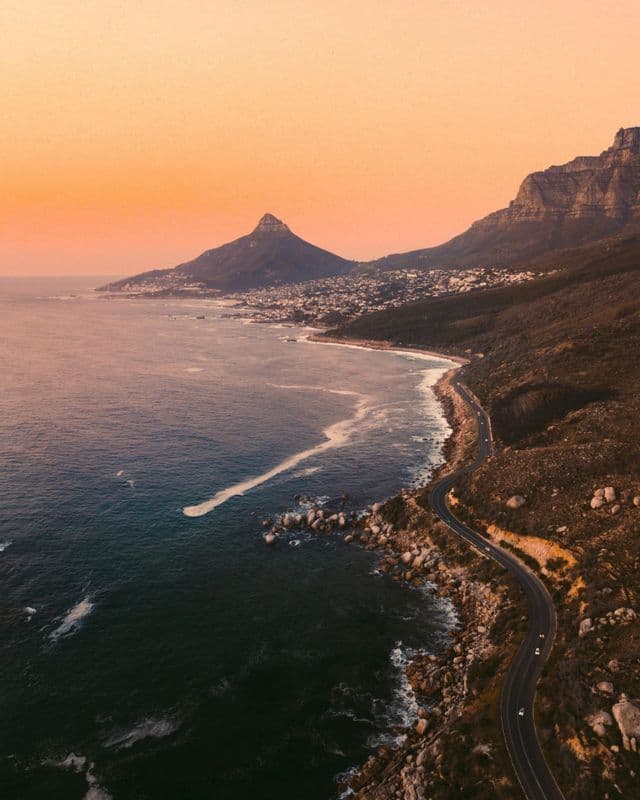 Una vista aerea di una strada tortuosa lungo una costa rocciosa, con una montagna e una città costiera in lontananza al tramonto.