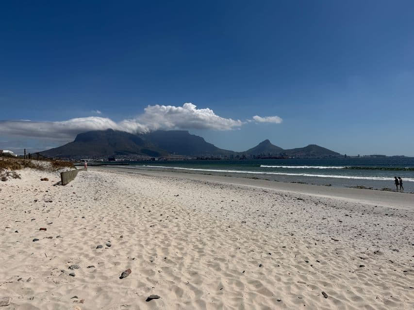 Vista da un'ampia spiaggia sabbiosa che si affaccia sull'acqua verso una grande catena montuosa dalla cima piatta, parzialmente coperta dalle nuvole.