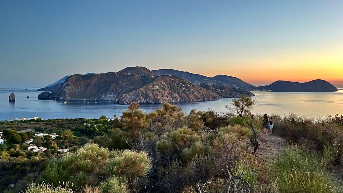 Due persone passeggiano su un sentiero panoramico in collina, affacciato su una baia con isole montuose e barche a vela al tramonto.