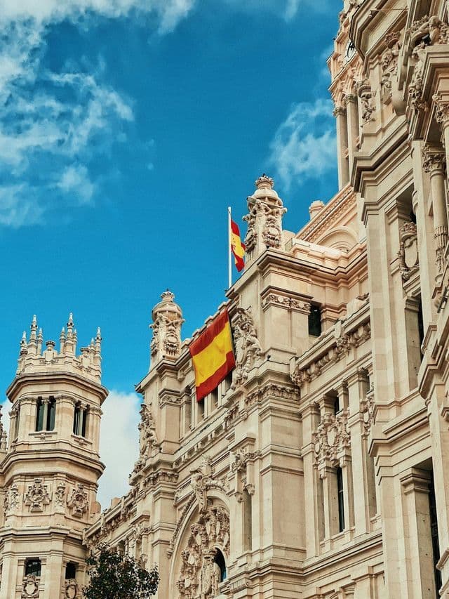 Spanish flags are displayed on the ornate facade of a historic building against a bright blue sky with white clouds.