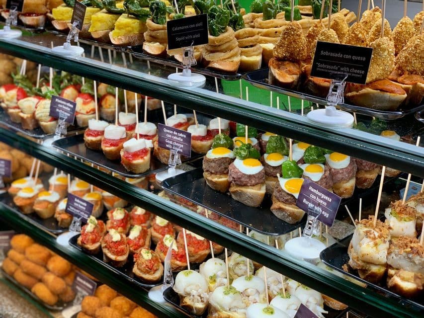 A variety of Spanish tapas, known as pintxos, neatly arranged on multiple shelves inside a glass display case.