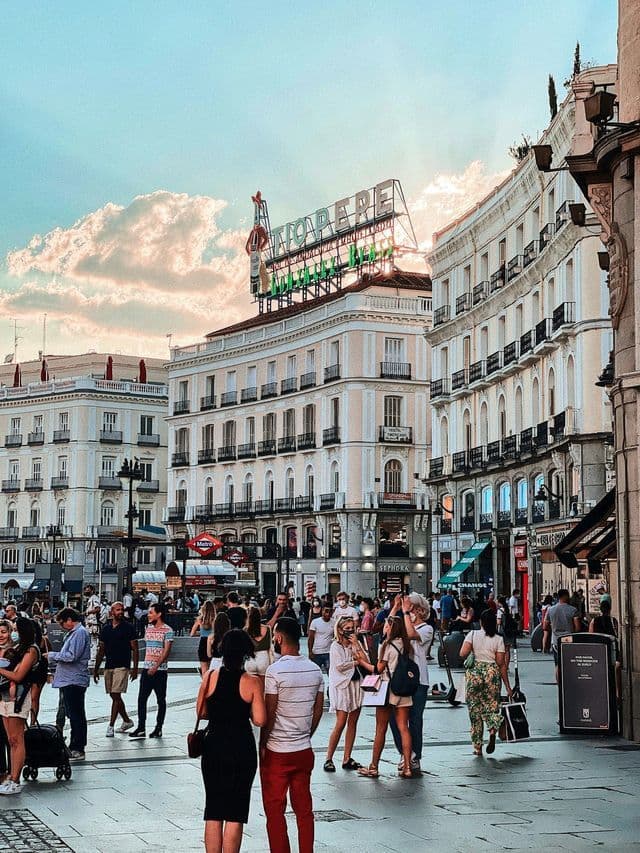 A crowd of people walking through a bustling city square, with classic European buildings and a large rooftop sign against a sunset sky.