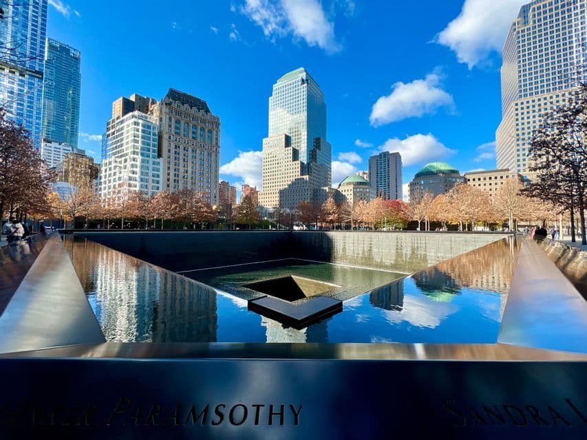 Una piscina reflectante conmemorativa con nombres grabados en su parapeto, que refleja los rascacielos circundantes de la ciudad y un cielo azul brillante.