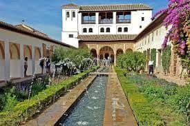 Un largo canal de agua fluye a través de un jardín de patio formal flanqueado por arcadas, con un palacio blanco al fondo.