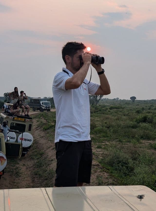 Un hombre en un viaje en grupo de WeRoad mira a través de binoculares la puesta de sol durante un safari en la sabana.