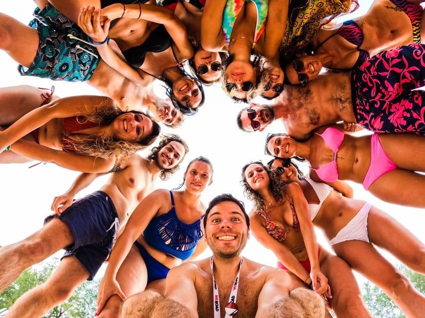 Un grupo de viaje de WeRoad en bañador se reúne en círculo, sonriendo para una selfie a contrapicado bajo un cielo brillante.