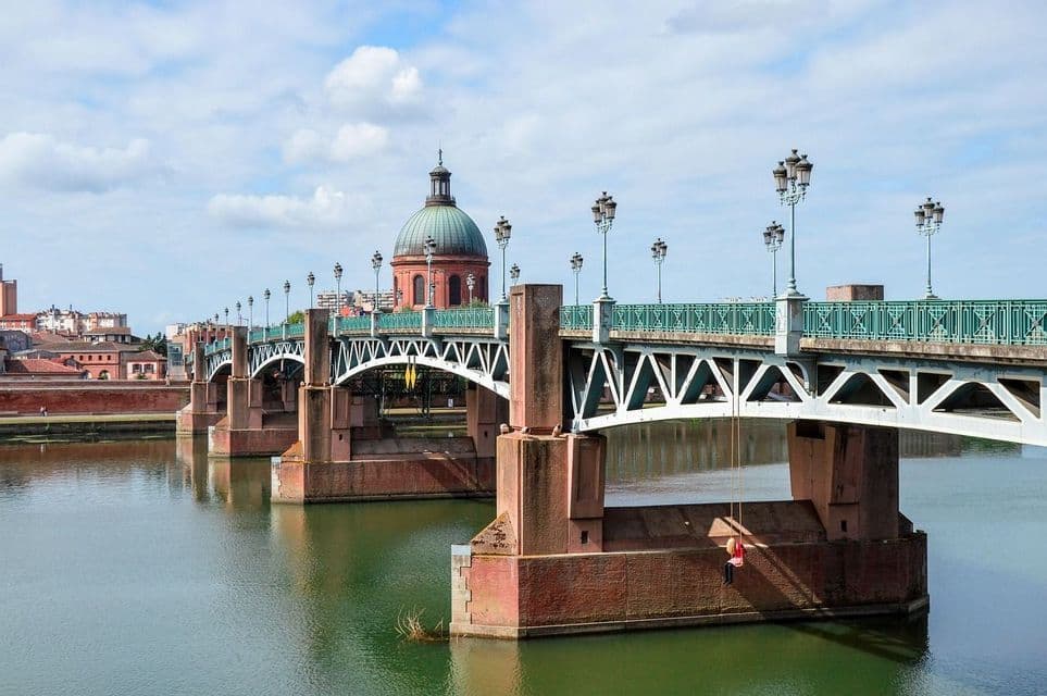 Una persona seduta su un'altalena di corda, appesa a un grande ponte su un fiume, con lo skyline di una città e un edificio a cupola sullo sfondo.